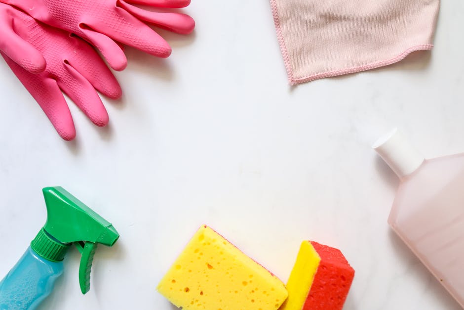 A flat lay of cleaning supplies on a white surface, including a pair of pink rubber gloves, a pink cloth, a green spray bottle, a yellow and red cleaning sponge, and a pink spray bottle, arranged to illustrate tools used for surface cleaning and sanitisation in domestic cleaning. The setup emphasizes attention to cleanliness, with the items neatly organized and ready for use by Cleaners Putney as part of a deep clean for Upper Richmond Road homes, highlighting effective hygiene and maintenance practices.
