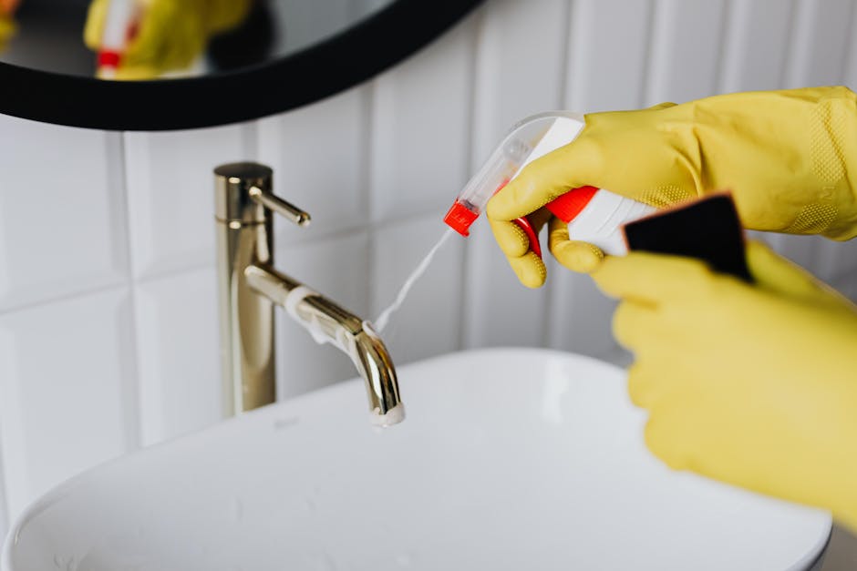 Close-up view of a person wearing yellow rubber gloves applying a liquid cleaning spray from a spray bottle onto the surface of a white ceramic sink in a bathroom. The sink has a modern, stainless steel faucet, and the background features white tiled walls with a black circular mirror. The area appears clean and well-maintained, demonstrating effective surface cleaning and hygiene practices as part of domestic cleaning services offered by Cleaners Putney, aligned with the deep clean guide for Upper Richmond Road homes.