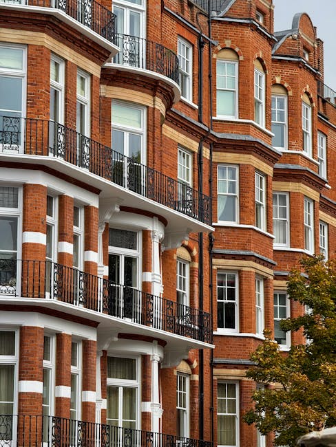 Photograph of a multi-story Victorian residential building with red brick façade, featuring white decorative trims and black wrought iron balconies. The windows are large with white frames, and the balconies have glass doors leading to the interior. The building is illuminated by natural daylight, highlighting the clean, well-maintained exterior. Nearby, a leafy tree adds greenery to the scene. The image is associated with Cleaners Putney, a company specializing in surface cleaning and deep cleaning services, including mould and damp cleanup for Victorian homes in Putney, supporting hygienic maintenance and preservation of such residential properties.
