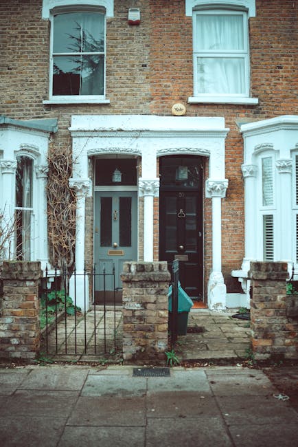 A photograph of two adjacent Victorian terraced houses in Putney, highlighting their brick facades and white-painted front entrances. The house on the left features a grey door with a glass panel, complemented by white decorative trim around the doorway and twin windows with white frames and curtains on the upper floor. In front of both houses, there are small gardens bordered by low brick walls and metal railings. The garden surfaces are paved with concrete slabs, with some patches of overgrown greenery and a narrow pathway leading to each entrance. Visible cleaning tools such as a doormat and a small outdoor light are present, indicating maintenance activities. The image captures a well-maintained residential facade in need of routine cleaning and preservation, emphasizing the importance of surface cleaning and deep cleaning for Victorian homes, as promoted by Cleaners Putney.