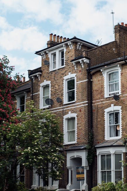 Photograph of a multi-story Victorian residential building with red brick façade, featuring white decorative trims and black wrought iron balconies. The windows are large with white frames, and the balconies have glass doors leading to the interior. The building is illuminated by natural daylight, highlighting the clean, well-maintained exterior. Nearby, a leafy tree adds greenery to the scene. The image is associated with Cleaners Putney, a company specializing in surface cleaning and deep cleaning services, including mould and damp cleanup for Victorian homes in Putney, supporting hygienic maintenance and preservation of such residential properties.