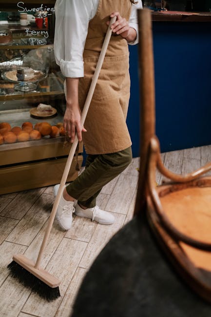 A person wearing a white shirt, beige apron, and dark pants is seen sweeping the wooden floor of a bakery or café with a long-handled broom. The individual is standing near a display case filled with
