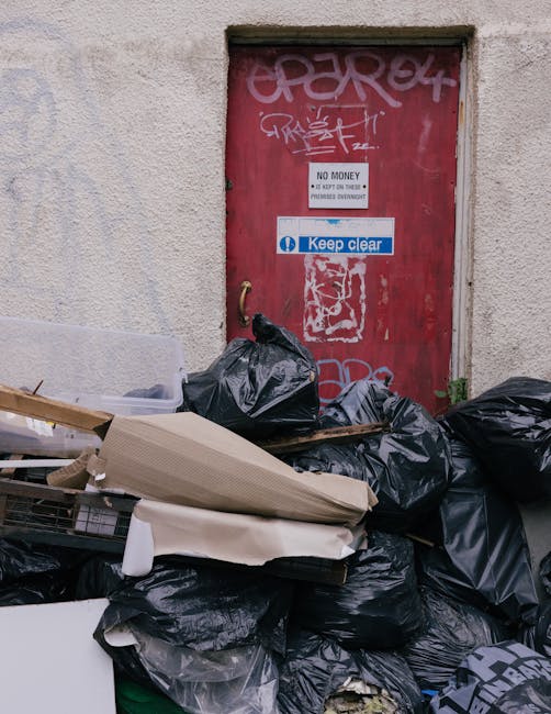 The image shows a cluttered outdoor space in front of a red metal door with graffiti and stickers, including a prominent white and blue 'Keep Clear' sign and a 'No Money' notice. The area is filled with black garbage bags, some partially open revealing discarded clutter, and discarded cardboard and paper packaging, suggesting waste accumulation. The wall surrounding the door is light-colored and textured, with visible wear and dirt. The scene indicates unmanaged waste, emphasizing the need for proper rubbish disposal and cleaning, which is addressed by professional domestic cleaning services provided by Cleaners Putney. This visual context relates to maintaining cleanliness and hygiene in residential or commercial premises, aligning with your page about skip permit rules and fines in Putney, where proper waste management is crucial.