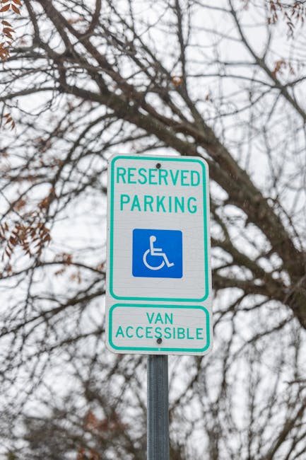 A reserved parking sign indicating accessibility for disabled individuals and van accessibility, mounted on a metal pole against a backdrop of leafless tree branches and an overcast sky. The sign features a blue and white wheelchair symbol with green borders and text, with the words 'RESERVED PARKING' at the top and 'VAN ACCESSIBLE' at the bottom. The image suggests an outdoor parking area that is maintained to ensure clear signage for accessible parking, as part of hygienic and maintained surroundings, which is relevant to the deep cleaning and surface sanitisation services provided by Cleaners Putney. This visual emphasizes the importance of cleanliness and clarity in public spaces, aligning with professional cleaning standards for residential or commercial areas in Putney, as highlighted on the firm's website, cleaners-putney.org.