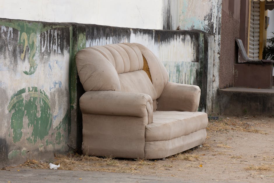 A worn beige upholstered armchair with visible tears and stains is placed outdoors on a dirt ground next to a weathered wall with peeling paint and graffiti. The chair has cushioned armrests and a high backrest, showing signs of neglect, with the fabric appearing flat and dirty. The surrounding environment includes a concrete step nearby and a partially visible building entrance with a louvered window, suggesting an urban setting. Natural light illuminates the scene, highlighting the worn condition of the furniture and the rough texture of the wall, reflecting the importance of proper furniture disposal and maintenance in line with Wandsworth Council rules on mattress and upholstery disposal, as promoted by Cleaners Putney for effective domestic cleaning and hygiene.
