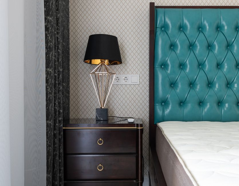 A close-up image of a bedroom corner featuring a dark wooden nightstand with two drawers, each with a circular gold handle. On top of the nightstand is a modern table lamp with a geometric gold wireframe base, black lampshade, and a gold interior that reflects light. The wall behind has textured wallpaper with a diamond pattern in neutral tones, alongside a white power outlet with multiple sockets. To the right, part of a bed with a tall, teal, tufted headboard with a glossy finish is visible, and a white mattress rests on a base beneath it. The room is well-lit, highlighting the sleek, clean surfaces and a minimalist, contemporary decor style. Cleaners Putney specializes in surface and domestic cleaning, ensuring hygiene and tidiness in residential spaces.